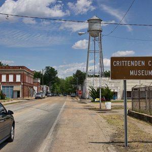 Street with parked cars town buildings sign with an arrow saying "Crittenden County Museum" and water tower