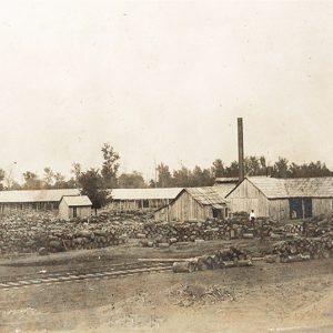 Large building complex with smokestack and cut timber covering the ground in front
