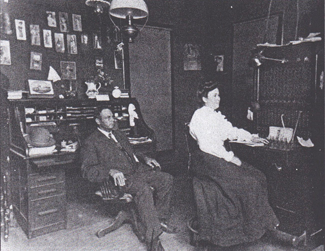 White man and woman at their desks in office with photographs on wall behind them