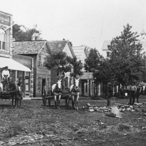 en and horse drawn wagons on dirt road with trees and brick and woodframe buildings behind them