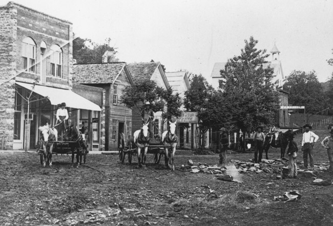 en and horse drawn wagons on dirt road with trees and brick and woodframe buildings behind them