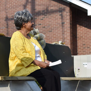Older African-American woman in yellow top white shirt and black pants sitting on bench outside brick building