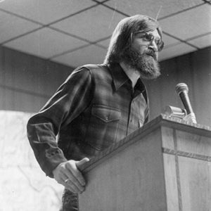 White man with bushy beard wearing glasses and plaid shirt speaking at lectern