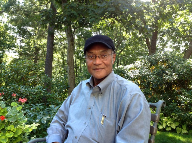 African-American man with glasses wearing a blue cap and button-up shirt sitting outside with trees and plants behind him