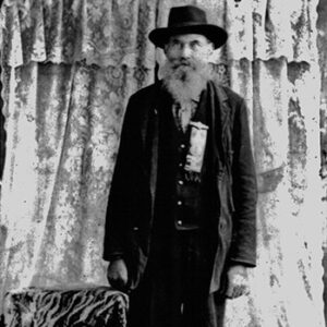 Older white man in hat and suit with long beard standing with curtains behind him