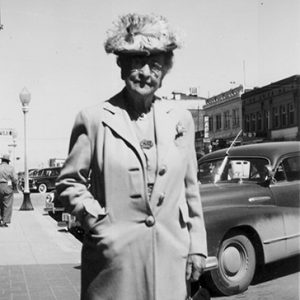 Old white woman in hat and long coat standing on street corner