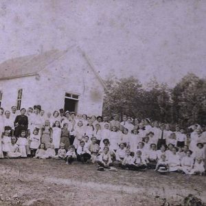 Congregation of white men women and children posing together outside single-story building