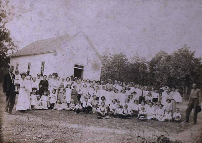 Congregation of white men women and children posing together outside single-story building