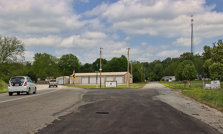 Street intersection with cars and metal garage building with outbuilding and radio tower in background