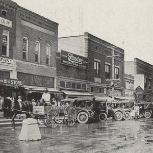 Horse drawn wagon and cars on street outside multistory brick storefront buildings