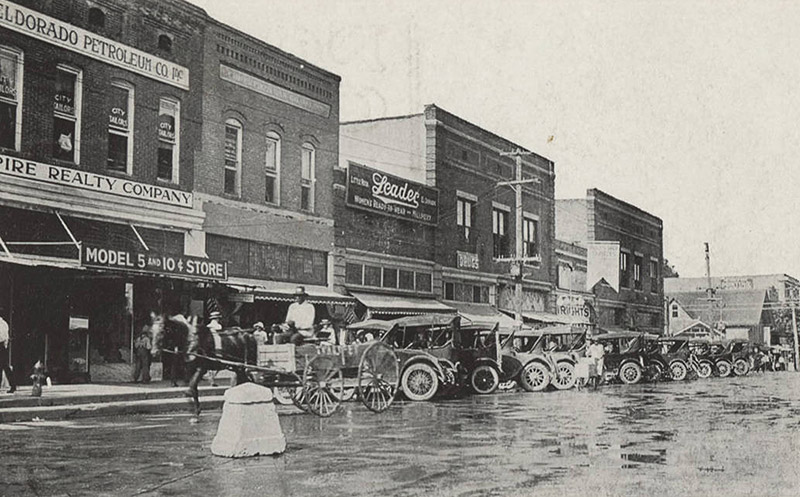 Horse drawn wagon and cars on street outside multistory brick storefront buildings