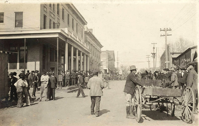 Crowded street outside multistory buildings with crowd of men and horse drawn wagon in the foreground