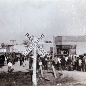 Crowded street with brick storefronts in the background and railroad crossing sign in the foreground