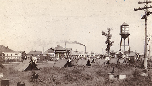 Men standing near rows of tents and water tower with town buildings in background
