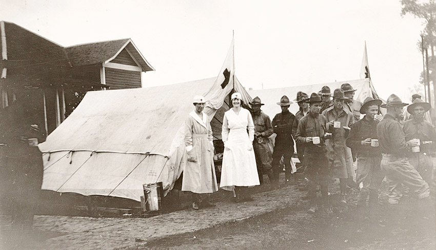 White soldiers with cups and two white women in nurses uniforms standing outside white tents