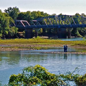 Two people standing in shallow river looking toward steel truss bridge and trees