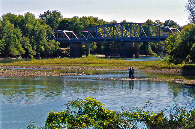 Two people standing in shallow river looking toward steel truss bridge and trees