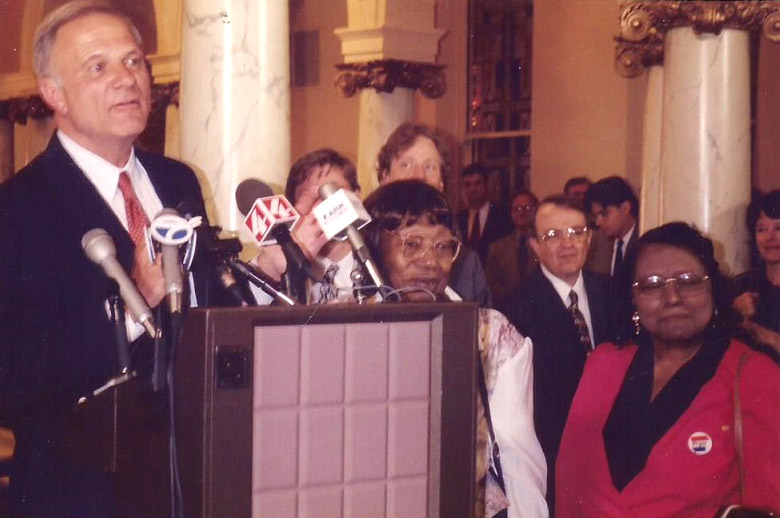 White man in suit speaking at lectern accompanied by African-American women with glasses