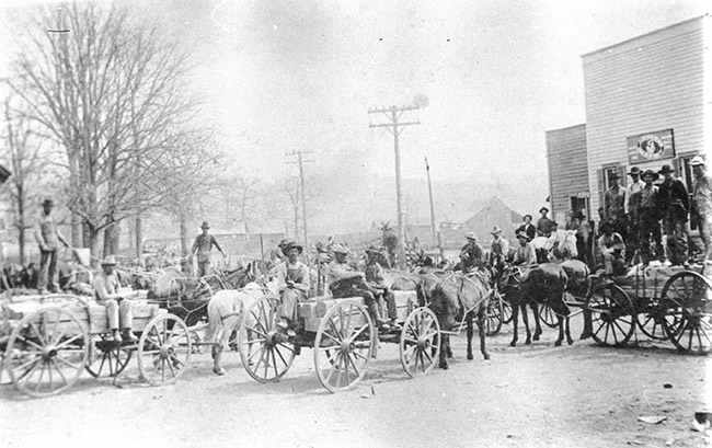 Crowded street with horse drawn wagons and white men in hats