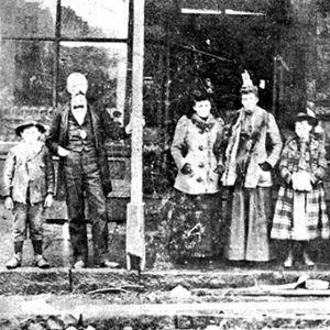 White men women and children standing outside storefront building with covered porch