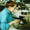 Older white woman with glasses in blue shirt making jewelry at her cluttered desk