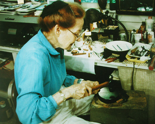 Older white woman with glasses in blue shirt making jewelry at her cluttered desk