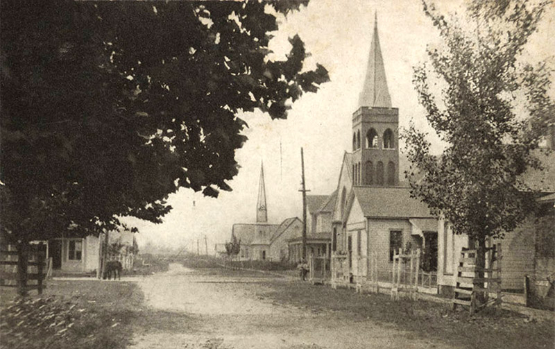 Two church buildings with bell towers and houses on town street with other buildings