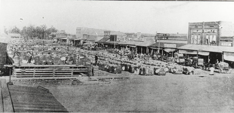 Bales of cotton in town square with row of storefronts on the right with horse drawn wagons in front of them