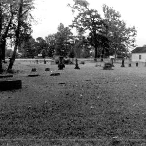 Stone monuments and gravestones in cemetery with single story building in the background