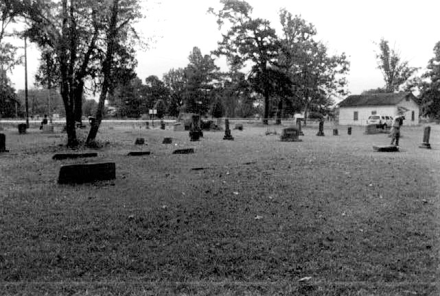 Stone monuments and gravestones in cemetery with single story building in the background