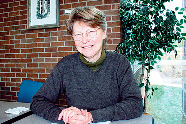 White woman in glasses wearing a sweater sitting at a table with brick wall behind her