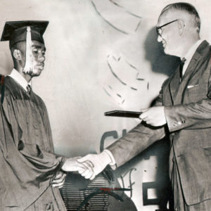 White man in glasses and suit shaking hands with young African-American man in cap and gown