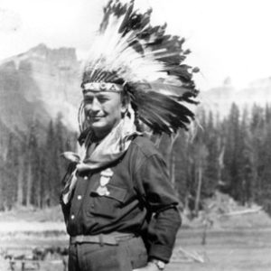 White man in feathered Native American head dress with trees and mountains in the background