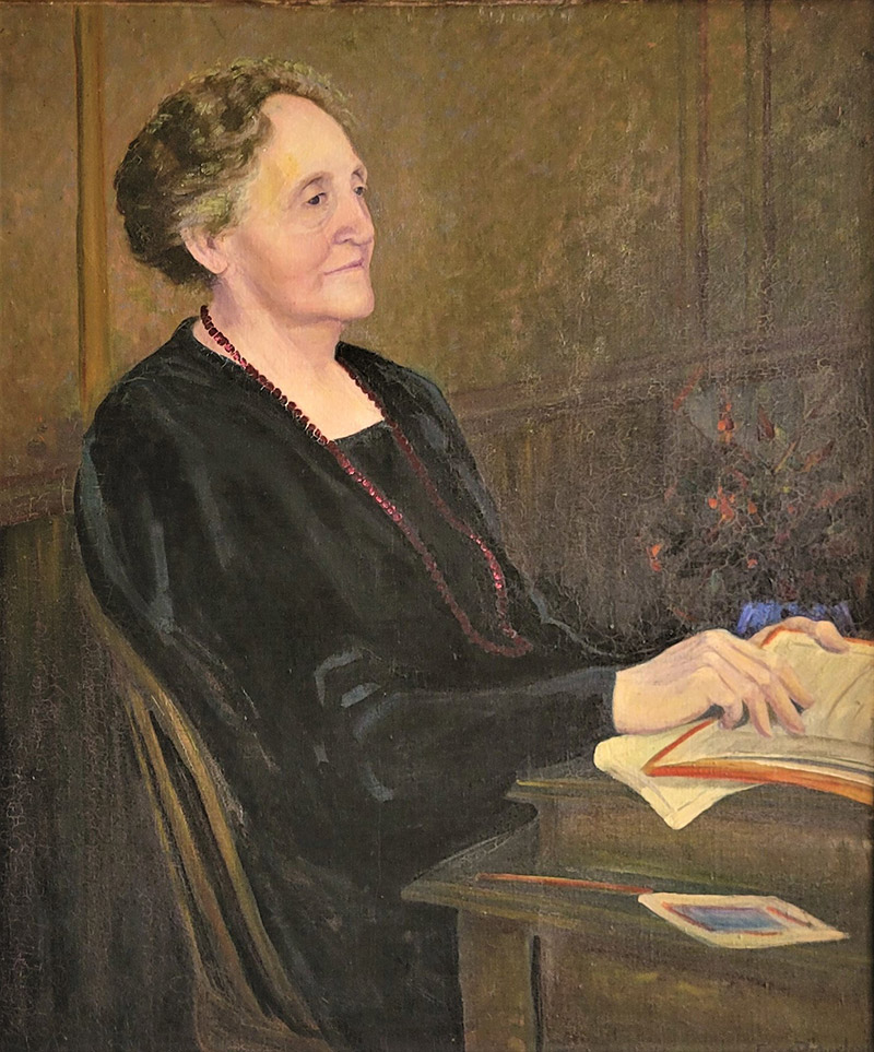 Older white woman sitting in black dress with red pearl necklace