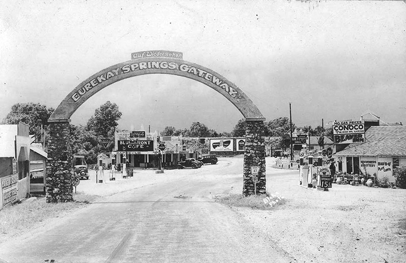 "Eure-Kay Springs Gateway" arch with rock columns over street with cafe and various businesses in the background