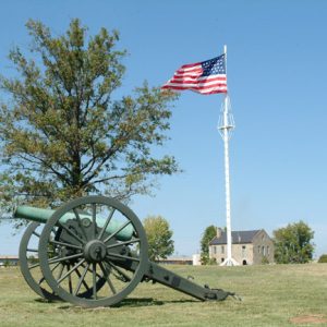 Large cannon on wheels in field with trees flag pole and multistory building in the background