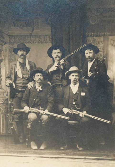 Five white men in western clothing and wide brimmed hats posing with rifles in hand