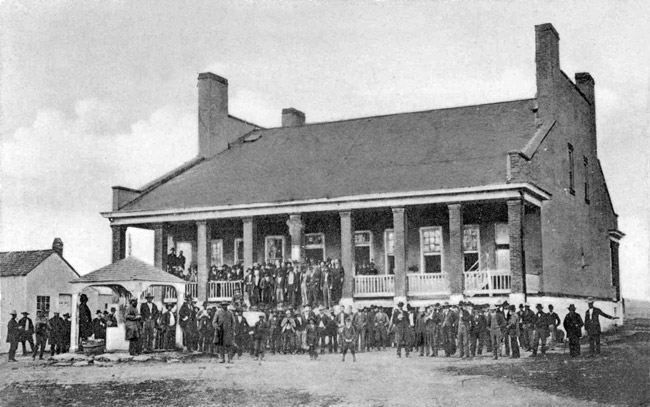 Crowd of white men in suits and hats standing outside of and on steps of two-story brick building with covered porch and gazebo