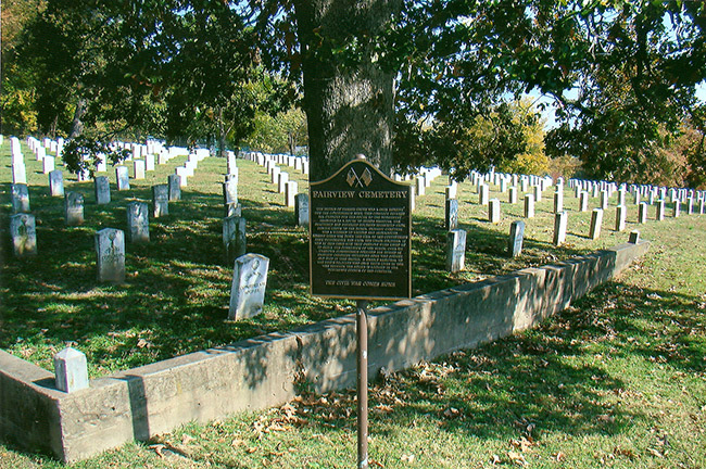 "Fairview Cemetery" sign next to wall and tree with rows of gravestones under it