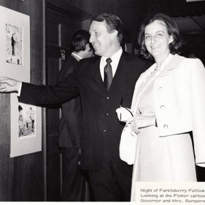 White man in suit and woman in dress smiling while looking at framed cartoons on wall beside them