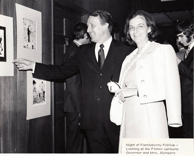White man in suit and woman in dress smiling while looking at framed cartoons on wall beside them