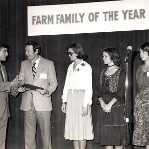 White man in suit handing award to white man in suit standing next to white women in dresses on stage under "Farm Family of the Year" banner