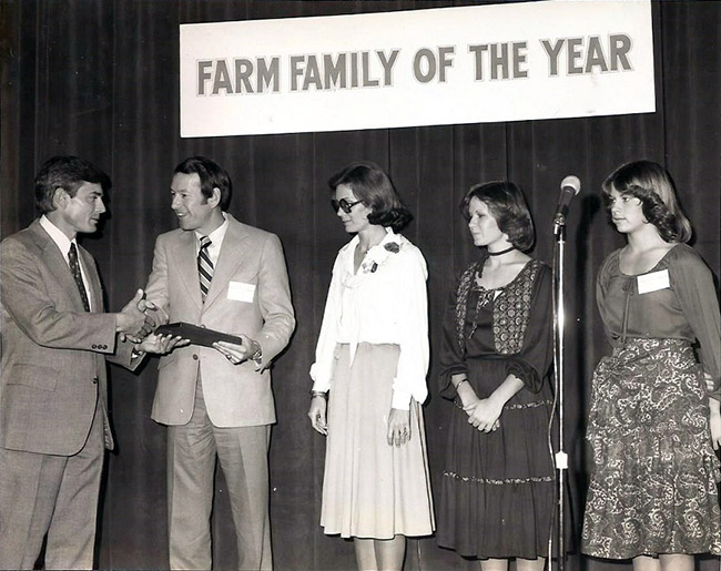 White man in suit handing award to white man in suit standing next to white women in dresses on stage under "Farm Family of the Year" banner