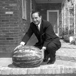 White man in suit and tie posing on a porch with a large watermelon