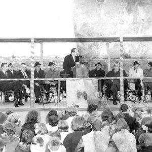 White man in suit speaking at lectern on raised platform with other white men in suits sitting behind him and large crowd before him
