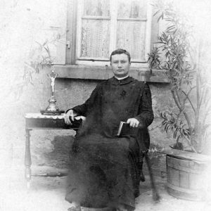White man in robes sitting with cross on table and plant