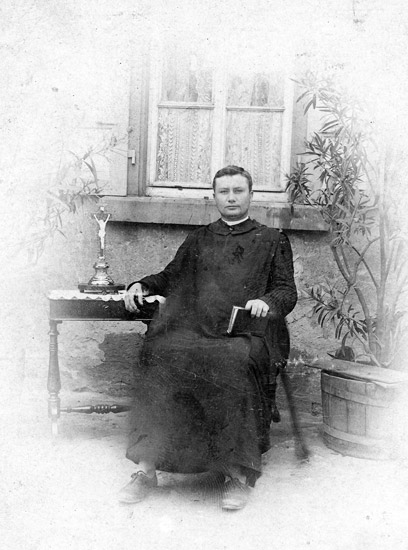 White man in robes sitting with cross on table and plant