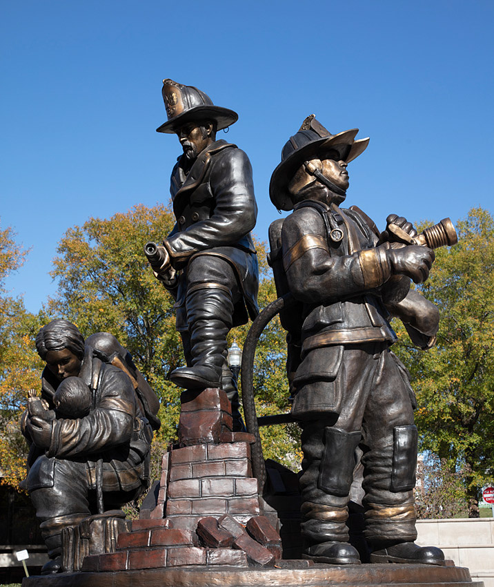 Bronze statue of two firefighters in full gear using hoses while a third firefighter comforts a small child behind them
