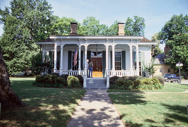 Single-story white house with brown wood doors covered porch and two brick chimneys