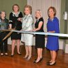Group of older white women indoors cutting a ribbon with large scissors